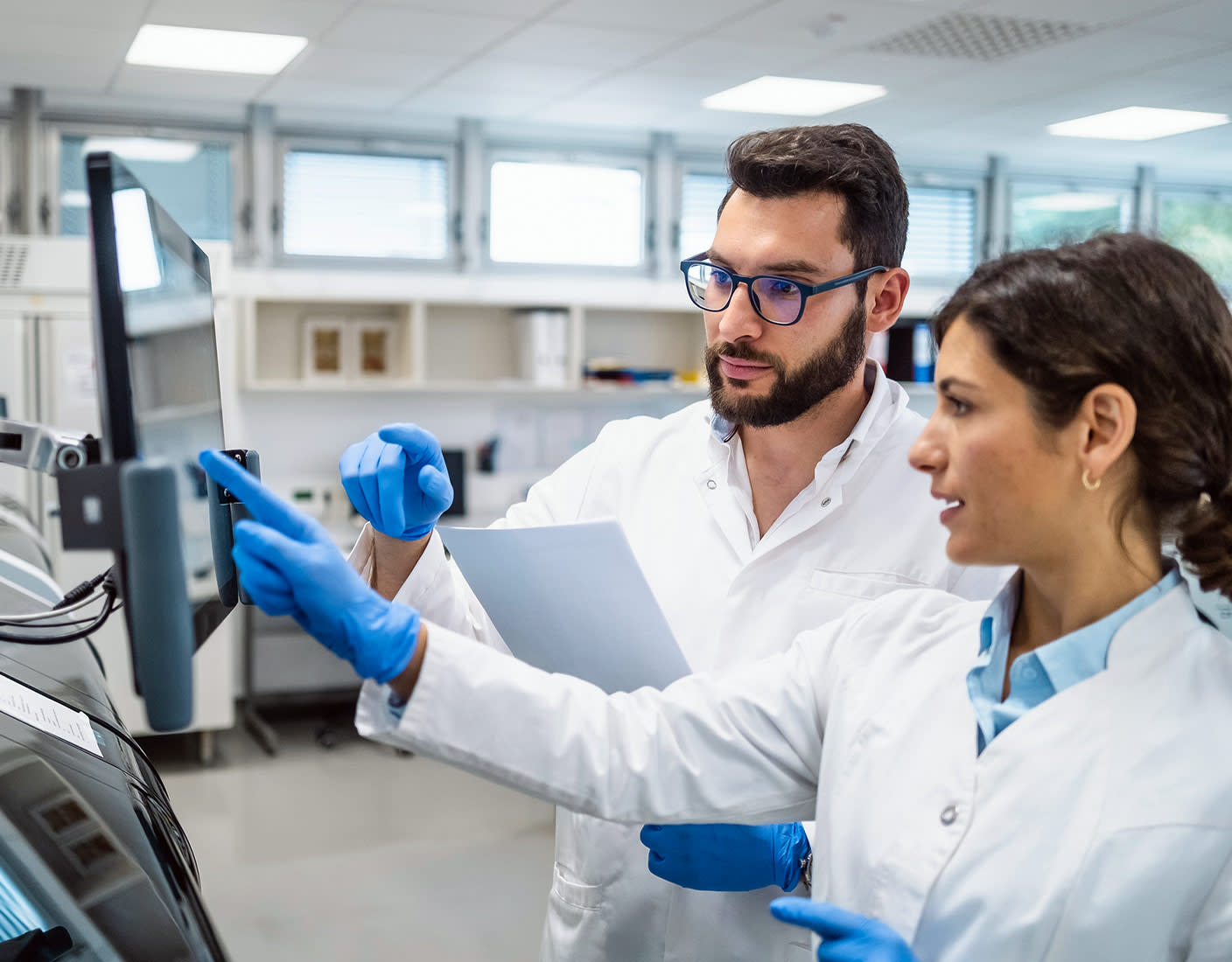 Two scientists in lab coats and blue gloves examine data on a monitor in a laboratory. The woman points to the screen while the man holds a document.